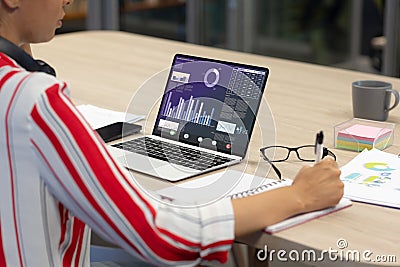 Midsection of asian businesswoman making report while studying graphical data on laptop in office Stock Photo