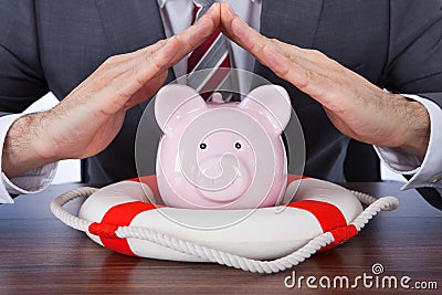 Businessman Sheltering Piggybank With Lifebelt At Desk Stock Photo