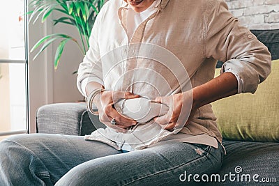 Midsection of woman hands holding her belly fat sitting on sofa at home. Caucasian lady grabbing excessive fat on her abdomen. Stock Photo