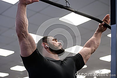 Mighty strong caucasian male with black beard wearing wireless headphones holding crossbar of exercise machine preparing for Stock Photo