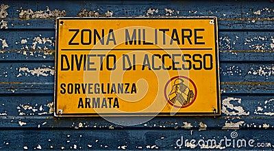 Military area with yellow sign: `Zona militare, divieto di accesso, sorveglianza armata`. Translation: `Milita Stock Photo