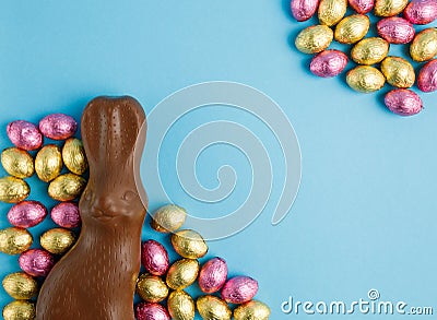 Milk chocolate Easter bunny and frame of pile of candy eggs wrapped in pink and golden foil on blue background Stock Photo