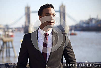 Millennial black businessman wearing black jacket, white shirt and tie standing at the Thames riverside in London, Tower Bridge in Stock Photo