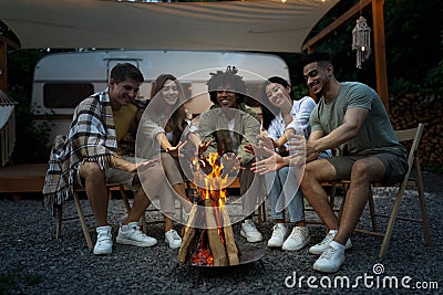 Millennial diverse friends sitting near bonfire, warming hands, having fun time together at night on camping trip Stock Photo