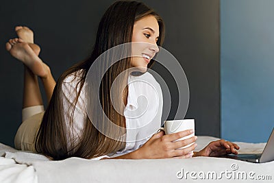 Millennial girl is resting on bed in white shirt, holding cup of coffee in her hand. Stock Photo