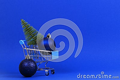 Mini shopping cart with small Christmas tree and navy Christmas decoration bauble balls on blue background Stock Photo