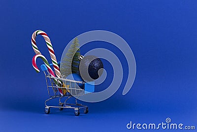 Mini shopping cart with small Christmas tree, Xmas decoration bauble balls and candy canes on navy blue background Stock Photo