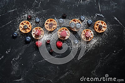 Mini waffles with raspberries and blueberries on a black stone background, flat lay. Belgian traditional waffles with berries. Top Stock Photo