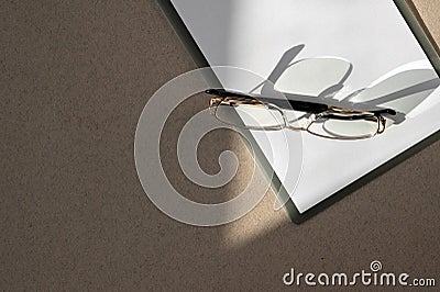 Minimal business brand template. Clipboard with white paper sheet closeup, eyeglasses on brown table with natural sunlight shadows Stock Photo