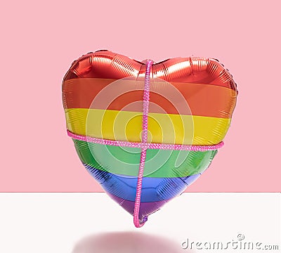 Minimal tight love concept. Rainbow color heart-shaped balloon with pink sparkle rope crosses it. Pink and star white background Stock Photo