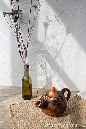 Minimalist still life with crafted brown clay teapot, bottle with flowers on wooden table, aesthetic sun light shadows on white Stock Photo