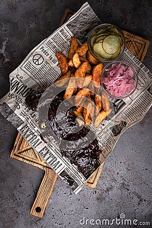 Minsk, Belarus - August, 2019 Top view on BBQ Pork ribs set with baked potato and pickled cucumber serve with newspaper on wooden Stock Photo