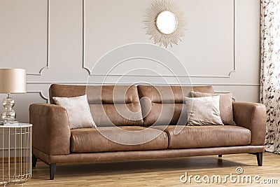 Mirror above leather couch with pillows in grey living room interior with gold lamp on table Stock Photo