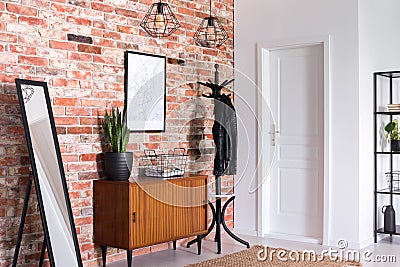 Mirror next to wooden cabinet in entrance hall interior with white door and poster on red brick wall Stock Photo