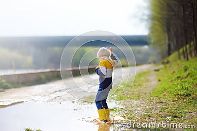 Mischievous preschooler child wearing yellow rain boots jumping in large wet mud puddle after rain. Kid playing and having fun in Stock Photo