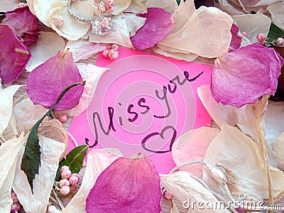 Miss you message on pink sticky note with dry rose and orchid flower petals and jewelry ring and chain on wooden background Stock Photo