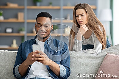 Mistrust. Attentive Lady Looking At Black Boyfriend While He Texting On Smarthone Stock Photo
