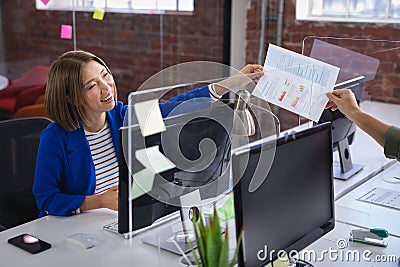 Mixed race female in front of computers separated by sneeze shield giving document to colleague Stock Photo