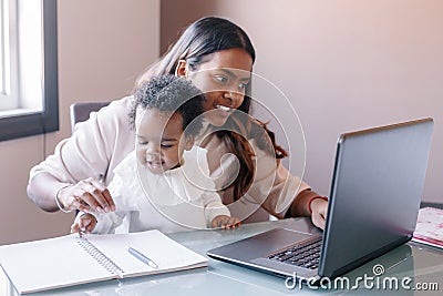 Mixed race Indian mother with African black baby working online from home on Internet. Workplace of freelancer woman with kid. Stock Photo