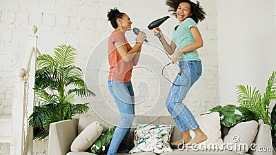 Mixed race young funny girls dance singing with hairdryer and comb jumping on sofa. Sisters having fun leisure in living Stock Photo