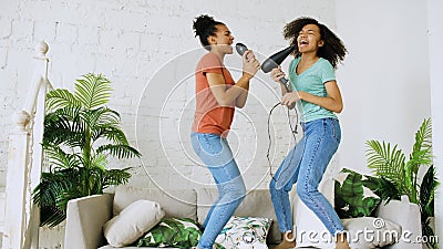 Mixed race young funny girls dance singing with hairdryer and comb jumping on sofa. Sisters having fun leisure in living Stock Photo