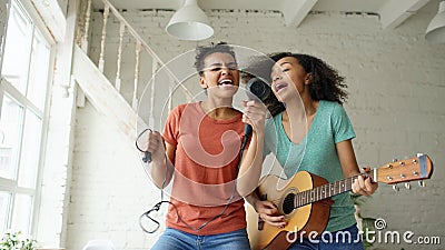 Mixed race young funny girls dance singing with hairdryer and playing acoustic guitar on a bed. Sisters having fun Stock Photo