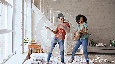 Mixed race young funny girls dancing singing and playing acoustic guitar on a bed. Sisters having fun leisure in bedroom Stock Photo