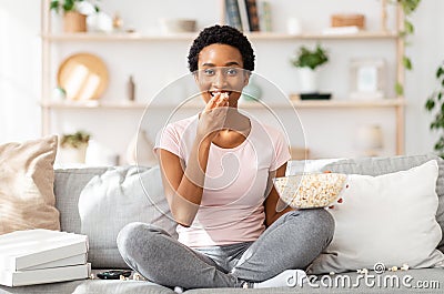 Mndless eating concept. Young black woman munching popcorn in front of TV on comfy sofa at home Stock Photo