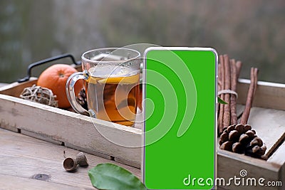 Mobile phone with green screen, glass of hot tea on tray, delicious vitamin tangerines with leaves, cinnamon sticks, pine cones, Stock Photo
