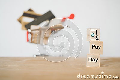 Mobile phone with magnifying glass and shopping cart icon on wooden cube block and pay day word, blurred credit cards in shopping Stock Photo