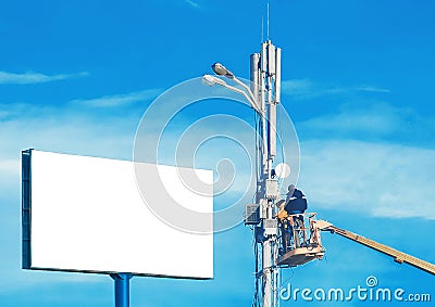 A mock-up large white billboard form and two construction workers on a machine crane repair a lighting pole against a blue sky Stock Photo