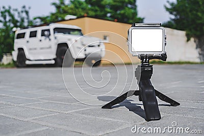 The mockup of the Action Camera stands on a tripod on paving slabs. Against the background of a white SUV car Stock Photo