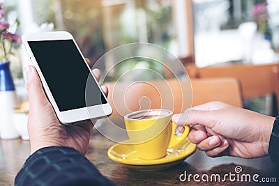 Mockup image of a hand holding white mobile phone with blank black desktop screen and yellow coffee cup on wooden table Stock Photo