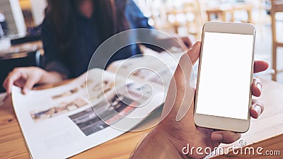 Mockup image of a man`s hand holding white mobile phone with blank screen in modern cafe and blur woman reading newspaper Stock Photo