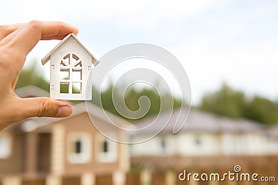 Model of white wooden house in hand against the background of cottages in the village. Dream of home, building, design, moving to Stock Photo