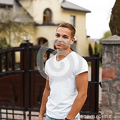 Modern American man with a fashionable hairstyle in a stylish white T-shirt stands near the wrought vintage gates near the house. Stock Photo