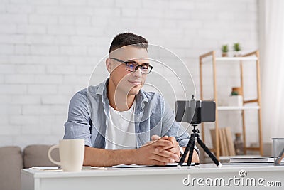 Modern blogger or teacher works from home. Young man looking at webcam, recording blog or webinar, uses smartphone with Stock Photo