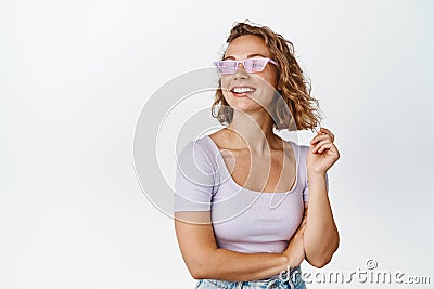 Modern blond girl in sunglasses, smiling and looking aside, wearing summer outfit, standing happy against white Stock Photo