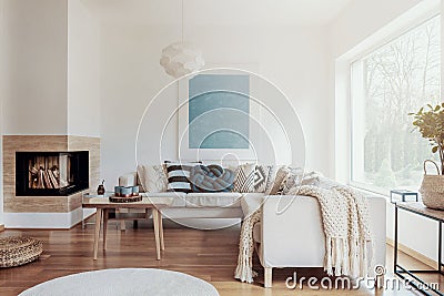 Modern corner fireplace in a sunny, peaceful living room interior with white walls and cozy pillows and blankets on a beige sofa. Stock Photo