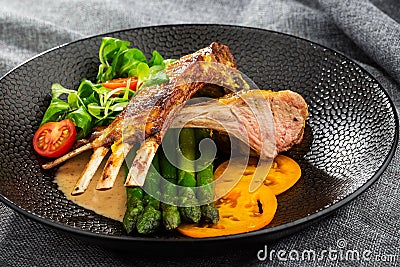 Roasted rack of lamb served with asparagus, yellow tomatoes and lamb sauce. Loin lamb Served in black stone plate with fork and Stock Photo