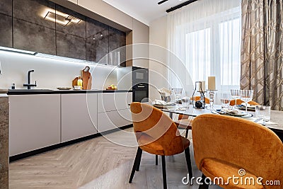 Modern kitchen combined with the dining room in dark and light colors, with orange chairs and glass cabinets for dishes Stock Photo