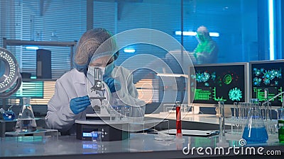 Modern medical research laboratory. Woman researcher in a white gown, mask, blue gloves and a bonnet is examining a Stock Photo