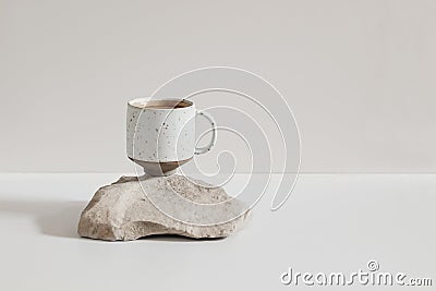 Modern minimal lifestyle still life scene. Speckled ceramic cup of coffee standing on sandstone rock, white table. Beige Stock Photo