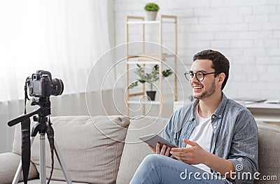 Modern profession is video blogging. Smiling guy with tablet in hands looks at camera and sits on sofa Stock Photo