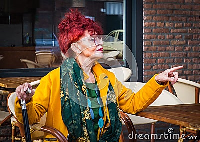 Fashionable woman in age sits in terrace of cafe and points with finger-modern lifestyle of pensionary, vacation or free time Stock Photo