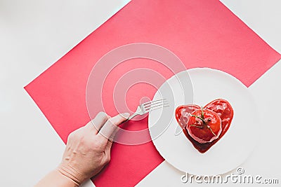 Modern still life scene. Fork in a female hand, tomato in heart shape tomato paste on a white plate. Stock Photo