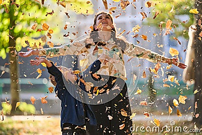 A modern woman joyfully plays with her son in the park, tossing leaves on a beautiful autumn day, capturing the essence Stock Photo