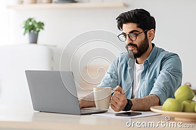 Modern worker in self-isolation. Concentrated arab man drinking coffee and working on laptop at table in kitchen Stock Photo