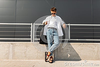 Modern young man hipster in fashionable summer white clothes in sunglasses in stylish red leather sandals with a bag stands Stock Photo