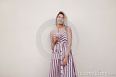 Modern young positive woman in a stylish long pink striped sundress stands near a vintage wall with a tasty milkshake in hands. Stock Photo